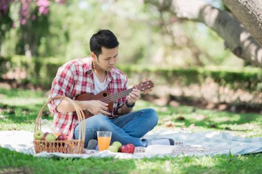 Kırmızı gömlekli bir adam ukulele oynuyor ve kitap okuyor, piknik konsepti.