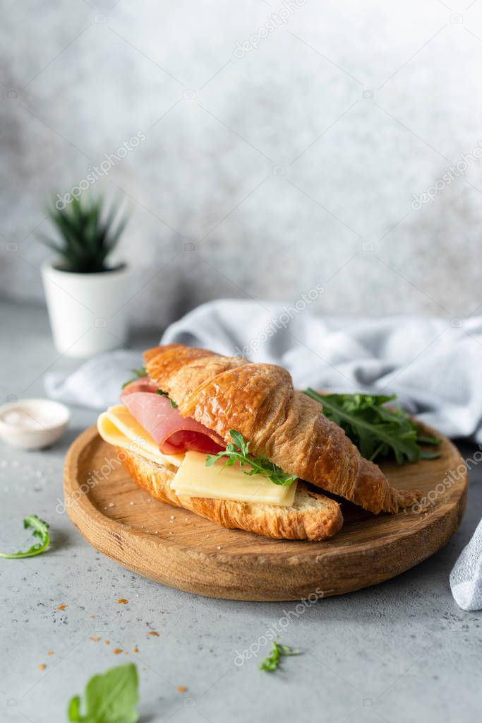 Croissant sandwich with cheese, ham and arugula on wooden cutting board, gray concrete background. Selective focus. Tasty breakfast sandwich or snack