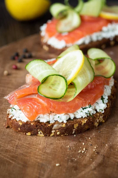 Tasty smoked salmon sandwich with cream cheese, cucumber and slice of lemon on wooden board, closeup view, selective focus