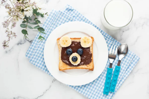 Food art bear toast for kids with chocolate nut spread, fruits and glass of milk. Top view on blue plaid towel