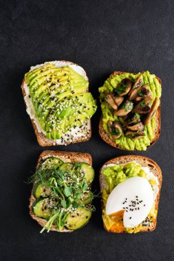 Siyah arka planda avokado tostları. Yumurtalı, salatalıklı, kızarmış mantarlı ve tohumlu avokado tostu.