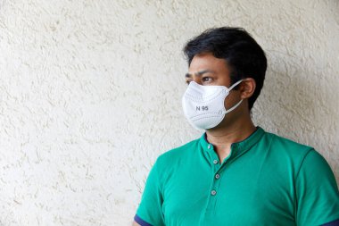 N95 beyaz solunum cihazı ve yeşil polo tişörtlü, kameranın soluna bakan Güney Asya kökenli yetişkin bir erkek resmi.