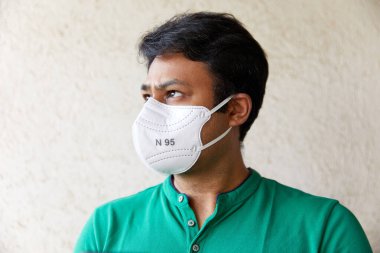 Yeşil polo tişörtlü beyaz N95 maskeli yetişkin bir adam kameraya bakıyor. Kafası 45 derece ve gözleri yukarı bakıyor.