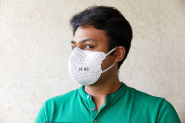 Yeşil tişörtlü beyaz N95 Solunum cihazı giyen yetişkin Hintli bir erkek kameraya bakıyor. Kafası 45 derece ve dokulu arka planı var.