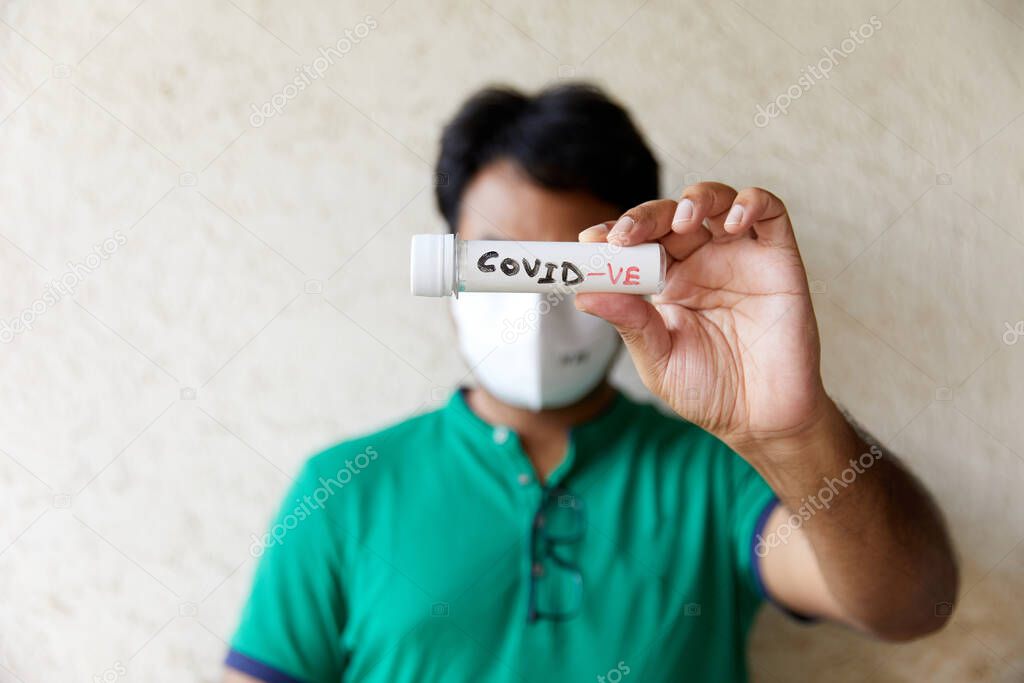 South Asian Male wearing a Green T-shirt holds a COVID Negative (COVID-ve) blood sample bottle in front of his face hiding the eyes. (Face Piece, White Mask, Half Face, Frontline Workers, Patient)