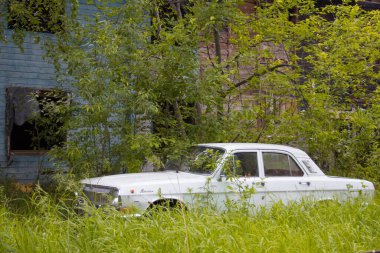Eski beyaz bir sedan, terk edilmiş bir ahşap evin yakınındaki, üzerinde uzun otlar olan bir otoparkta duruyor.