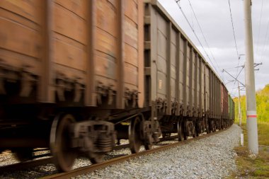 Bir yük treni raylar boyunca acele eder. Kahverengi gondol vagonları sütunları ve setleri süpürüyor.