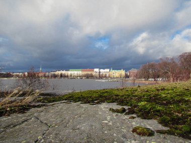 Helsinki 'deki binalar, ön planda kaya ve yosun bulunan bir adadan ve arka planda parçalı bulutlu bir gökyüzünden görülüyor..