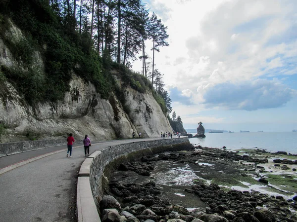 Stanley Park Vancouver 'da okyanus kıyısında koşucular ve Siwash Rock arka planda..
