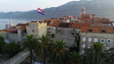 Korcula adasının ortasındaki kulede sallanan Hırvat bayrağı, 4K hava manzaralı. Günaydın, Korcula manzarası ve limana demirlemiş lüks yatlar ve yelkenliler..