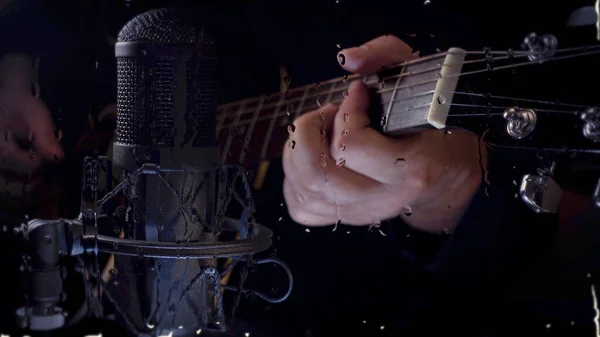 Musician plays guitar at studio near the microphone behind the glass with water drops