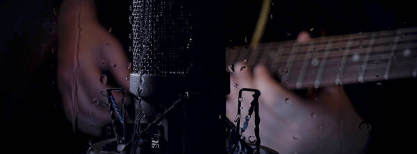 Музыкант играет на гитаре в студии возле микрофона за стеклом с каплями воды