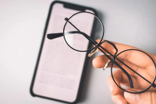 Hand holding reading glasses and a phone on a gray background. — Stock Photo, Image