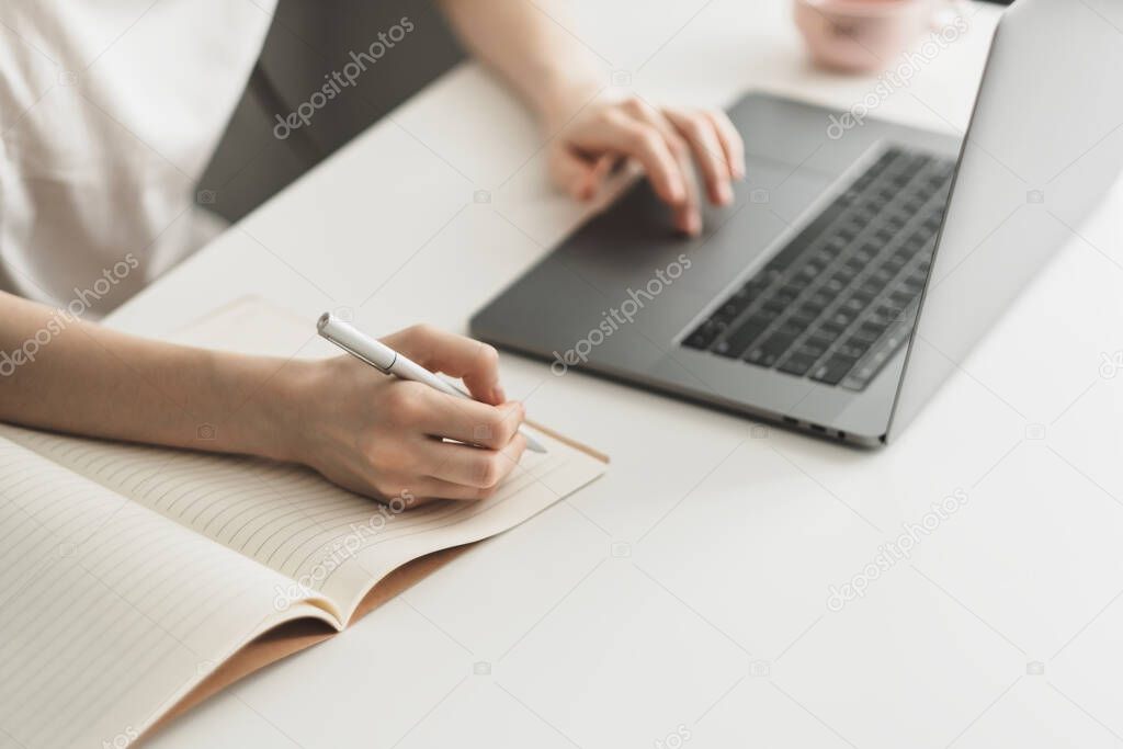 A young woman works on a laptop and takes notes in a notebook. 