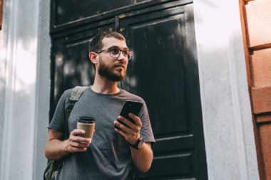 Sırt çantalı gülümseyen sakallı genç adam akıllı telefon kullanarak şehir caddesinde durup hazır kahve içerken. Yüksek kalite fotoğraf