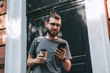 Sırt çantalı gülümseyen sakallı genç adam akıllı telefon kullanarak şehir caddesinde durup hazır kahve içerken. Yüksek kalite fotoğraf