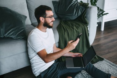 Gülümseyen genç sakallı adam, iş adamı akıllı telefon ve dizüstü bilgisayar kullanarak kanepenin yanında oturuyor. Evde çalış. Yüksek kalite fotoğraf