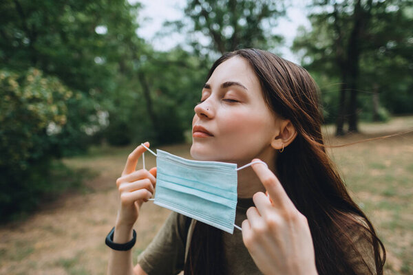 Young caucasian pretty woman removes a medical protective mask from her face on nature, surrounded by trees, breathing clean fresh air. High quality photo