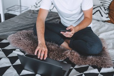 Yatakta oturan, akıllı telefon ve dizüstü bilgisayar kullanan serbest çalışan bir adam. Evde çalış. Yüksek kalite fotoğraf