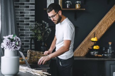Mutfak odasında dizüstü bilgisayar kullanan dalgın, beyaz sakallı genç bir adam. Yüksek kalite fotoğraf
