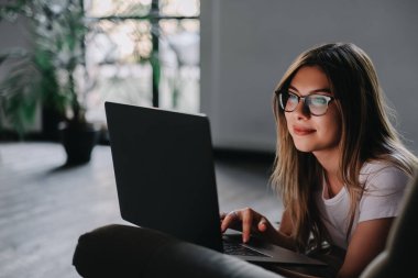 Kanepede dinlenen ve dizüstü bilgisayar kullanan güzel, genç, gülümseyen beyaz bir kadın. Arkadaşlarla sohbet etmek, bilgi taramak, online çalışmak. Yüksek kalite fotoğraf