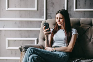 Kanepede dinlenen ve akıllı telefon kullanan güzel, genç, gülümseyen beyaz bir kadın. Arkadaşlarla sohbet etmek, bilgi taramak. Yüksek kalite fotoğraf