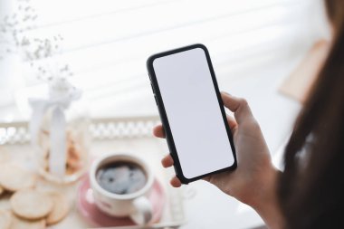 Kadın, sabahları pencerenin yanında beyaz ekranlı akıllı telefon kullanıyor. Model yap. Yüksek kalite fotoğraf