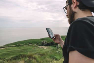 Genç sakallı beyaz adam güneşli bir günde deniz kenarında akıllı telefon kullanır. Yüksek kalite fotoğraf