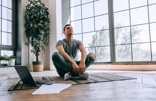 Pleased caucasian relaxed student sits crossed legs on floor near opened laptop, tries to have break after work. Yoga and meditation concept. High quality photo