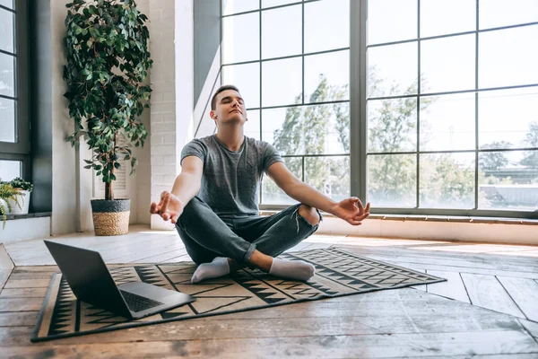 Pleased caucasian relaxed student sits crossed legs on floor near opened laptop, tries to have break after work. Yoga and meditation concept. High quality photo
