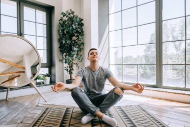 Kafkasyalı, rahat, kulaklıklı bir adam büyük pencerenin yanında bacak bacak üstüne atıyor ve işten sonra dinlenmeye çalışıyor. Yoga ve meditasyon konsepti. Yüksek kalite fotoğraf