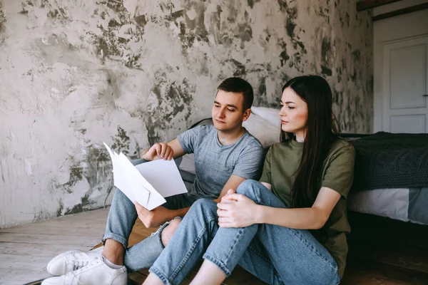 A young couple manages finances by looking through their bank accounts in the living room by the bed. Woman and man look at documents together. Planning budget expenses. High quality photo