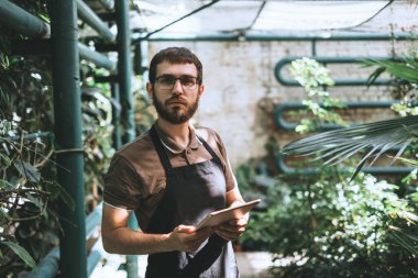 Genç bahçıvan gözlüklü ve önlüklü. Dijital tabletli. Daha iyi kalite kontrol için bahçe merkezinde çalışıyor. Çevreci serada dijital tablet kullanıyor. Yüksek kalite fotoğraf