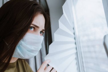 Tıbbi koruyucu maskeli beyaz bir kadın pencereden dışarı bakıyor. Coronovirus Karantina Konsepti. Yüksek kalite fotoğraf