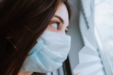 Tıbbi koruyucu maskeli beyaz bir kadın pencereden dışarı bakıyor. Coronovirus Karantina Konsepti. Yüksek kalite fotoğraf