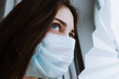 Tıbbi koruyucu maskeli beyaz bir kadın pencereden dışarı bakıyor. Coronovirus Karantina Konsepti. Yüksek kalite fotoğraf