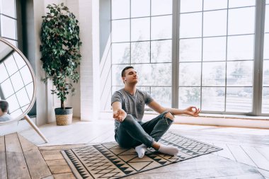 Kafkasyalı, rahat, kulaklıklı bir adam büyük pencerenin yanında bacak bacak üstüne atıyor ve işten sonra dinlenmeye çalışıyor. Yoga ve meditasyon konsepti. Yüksek kalite fotoğraf