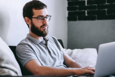 Gözlüklü genç bir adam dizüstü bilgisayarda çalışıyor, evdeki kanepede otururken internette geziniyor. Elektronik cihazlar üzerinde çalışan biri. Yüksek kalite fotoğraf