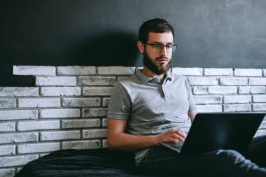 Gözlüklü genç bir adam dizüstü bilgisayarda çalışıyor, evdeki kanepede otururken internette geziniyor. Elektronik cihazlar üzerinde çalışan biri. Yüksek kalite fotoğraf