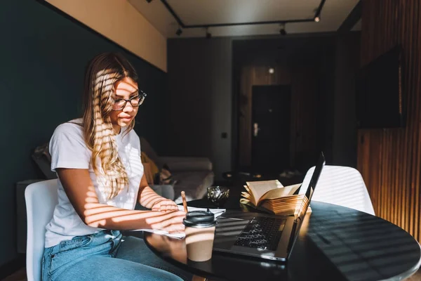 Focused caucasian young woman college student in glasses studying with books laptop distantly preparing for test exam writing essay doing homework at home, distantly education concept.