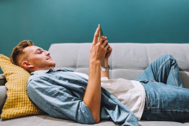 Genç beyaz adam evdeki kanepede uzanıyor ve tablet bilgisayar kullanıyor.