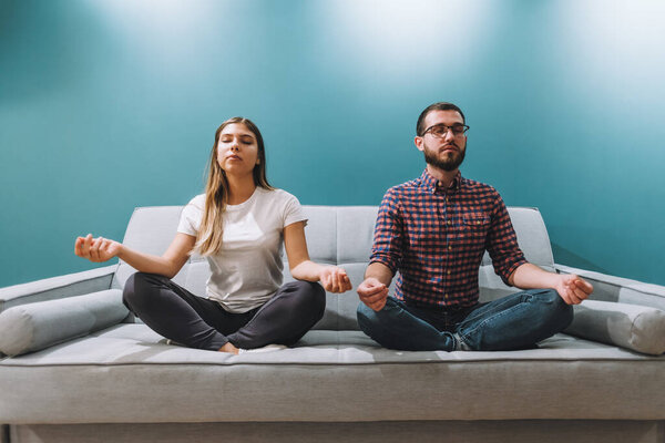 Young couple meditating together while sitting on sofa at home.