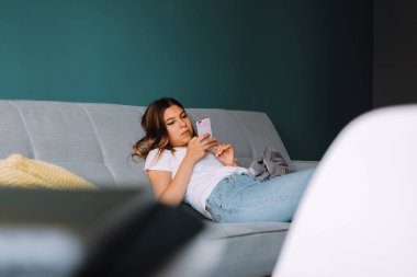 Oturma odasındaki kanepede dinlenirken akıllı telefon kullanan güzel bir genç kadın. Yüksek kalite fotoğraf