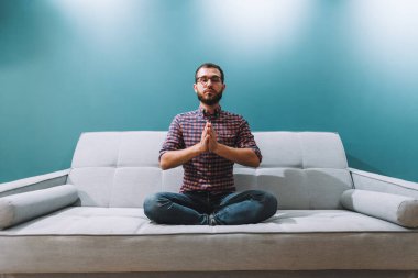 Gözleri kapalı genç bir beyaz adam kanepede oturuyor ve evde meditasyon yapıyor. Yüksek kalite fotoğraf