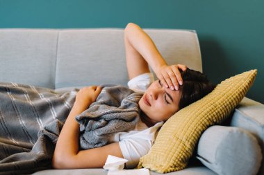 Alnına dokunan yorgun genç bir kadın baş ağrısı çekiyor, oturma odasındaki kanepede yatıyor. Yüksek kalite fotoğraf