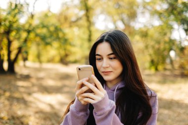 Akıllı telefon kullanan ve sonbahar havasının tadını çıkaran güzel bir genç kadın. Sonbahar parkında yürüyen bir kadın. Yüksek kalite fotoğraf