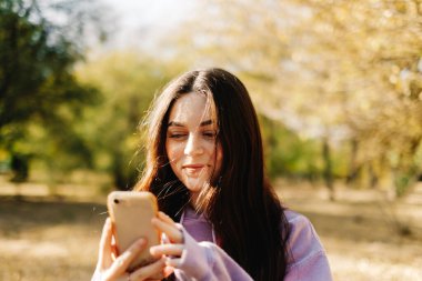 Akıllı telefon kullanan ve sonbahar havasının tadını çıkaran güzel bir genç kadın. Sonbahar parkında yürüyen bir kadın. Yüksek kalite fotoğraf
