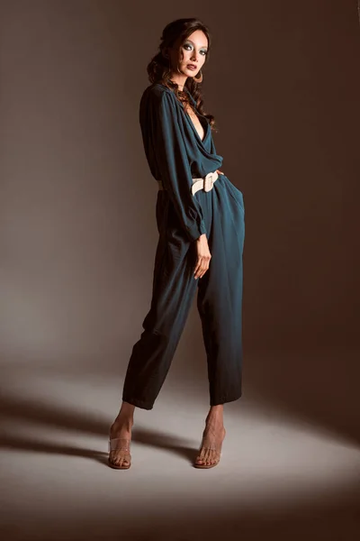 Esmer, genç bir kadın modellik yapıyor ve yeşil bir elbise giyiyor. Gri arka plandaki kameraya bakıyor.