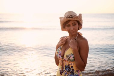 Issız bir plajın kıyısında duran Latin kız. Sarı mayo ve şapka giyiyordu. Şapkasına dokunuyor..