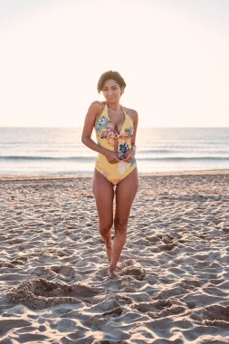 Latin kız, kumsalda yürürken sarı mayo giymiş kameraya bakıyordu.